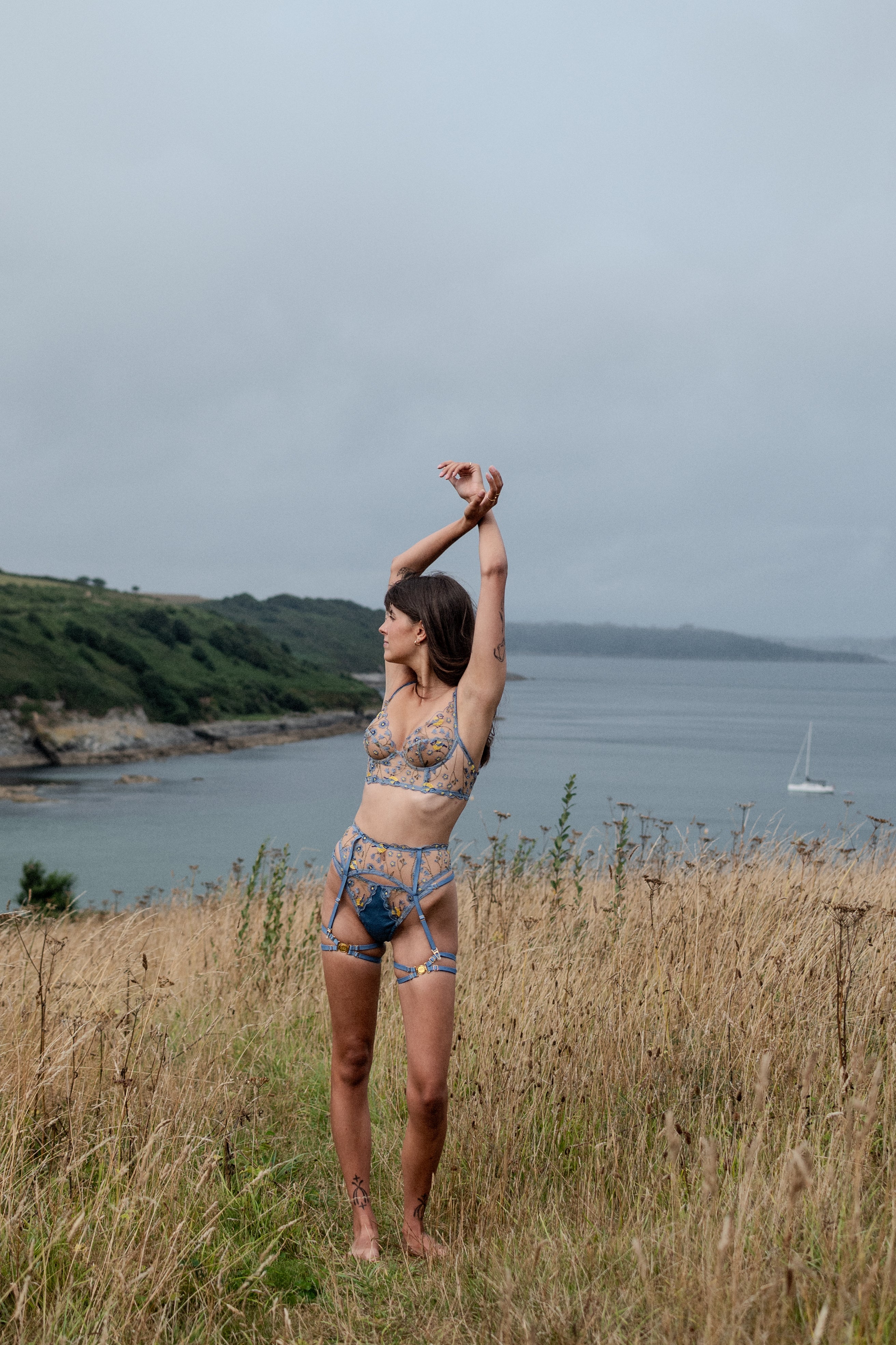 Model wearing blue floral embroidered plunge bra, suspender belt and g-string on cliff.